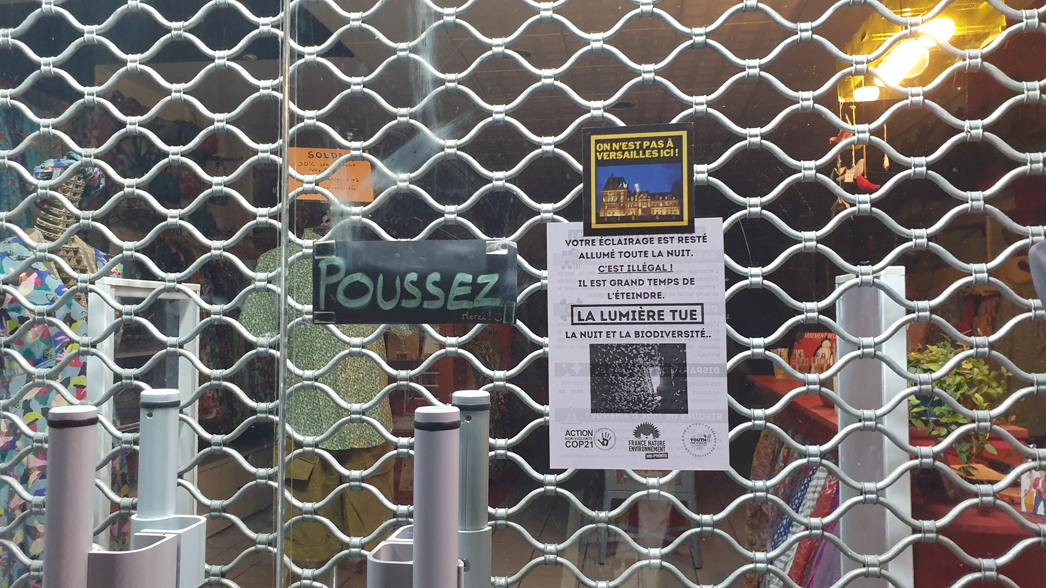 Grille métallique d'un commerce avec un sticker FNE affiché : « La lumière tue, la nuit et la biodiversité ».
