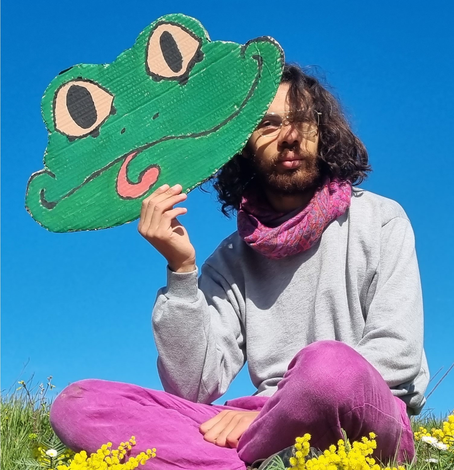 Portrait de Vie tenant un masque de grenouille devant son visage, assise dans un champ de mimosas, ciel bleu.